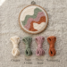 Wooden embroidery hoop containing abstract punch needle piece in cream, sage green, dusty rose, and terracotta, with four yarn threads displayed below labeled with color names